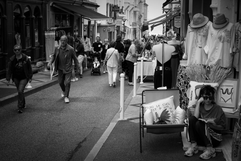 Una chica con gafas de sol lee un libro sentada en el suelo junto a un gato tumbado en una silla mientras la gente pasea por la calle, fotografía en blanco y negro