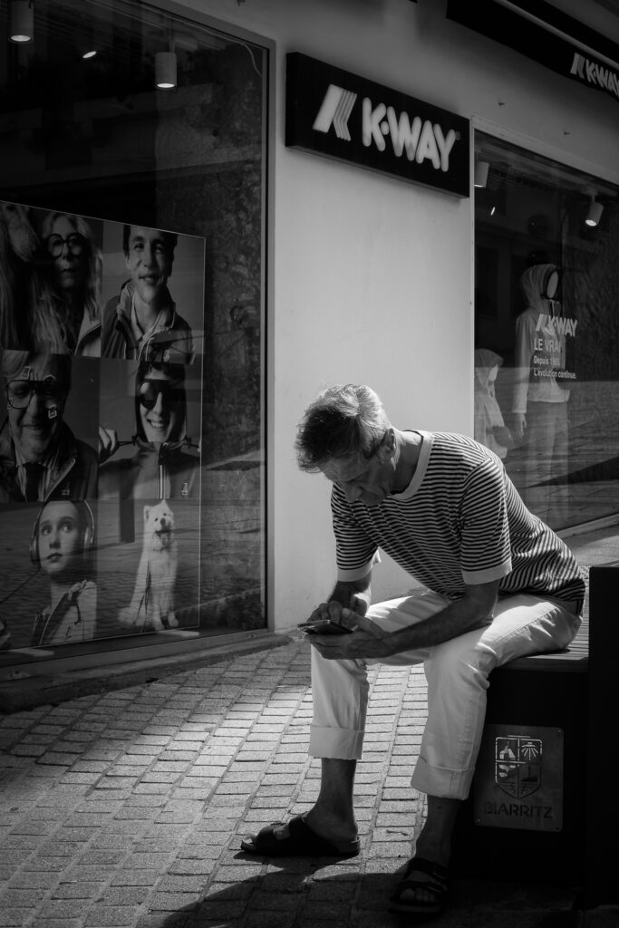 Hombre sentado sobre un banco con el escudo de Biarritz mirando su smartphone y escaparte de tienda a la izquierda, fotografía en Blanco y Negro