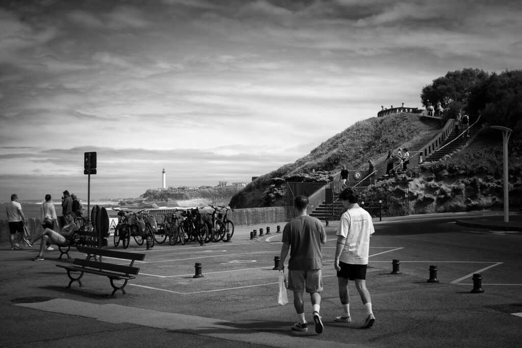 Dos chicos pasean con el faro de Biarritz al fondo cerca del acuario de Biarritz, fotografía en blanco y negro