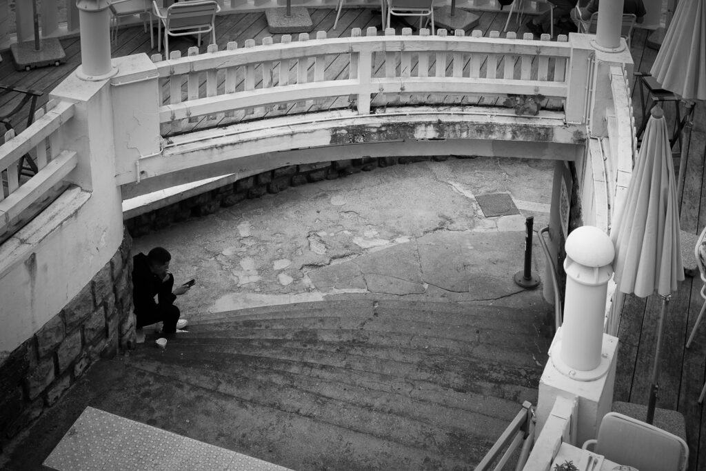 Un chico fuma sentado en unas escaleras bajo la terraza de un bar, fotografía en blanco y negro