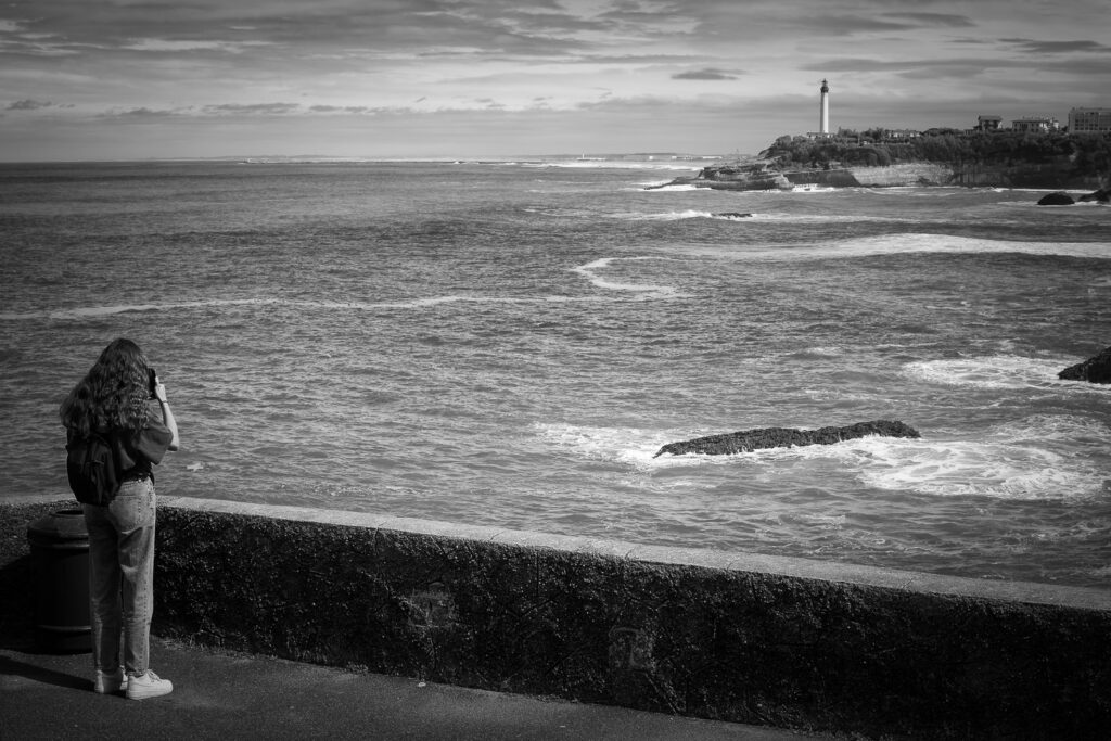 Una chica fotografía las vistas del mar con el faro de Biarritz al fondo, fotografía en blanco y negro