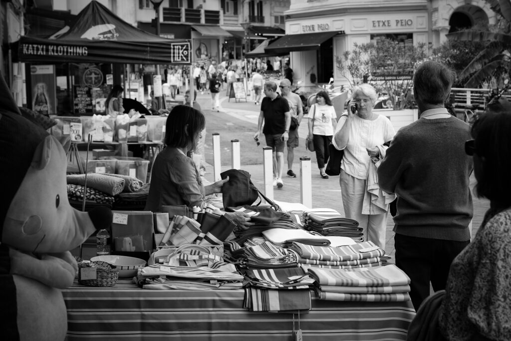 Una vendedora de un puesto del mercado ordena el género mientras los transeúntes pasean por la calle, fotografía en blanco y negro